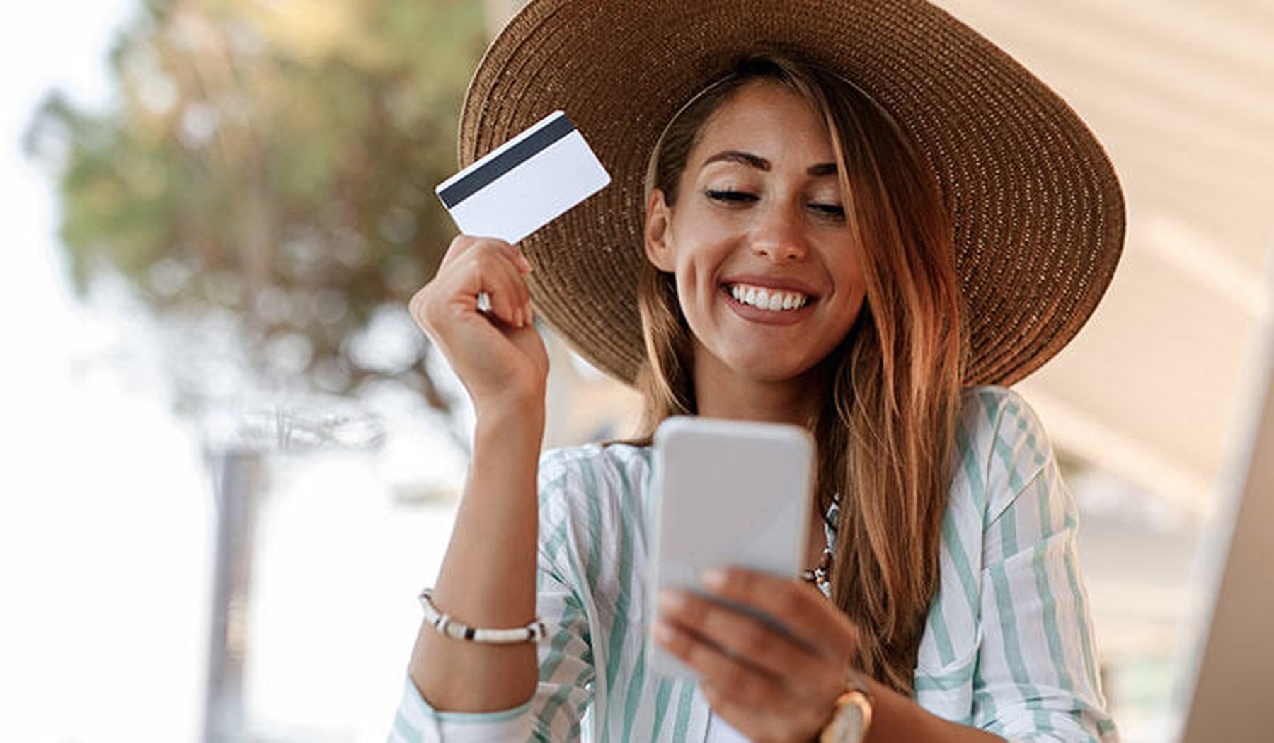 Een vrouw in een zomerse outfit houdt haar telefoon vast in de ene hand en haar bankkaart in de andere
