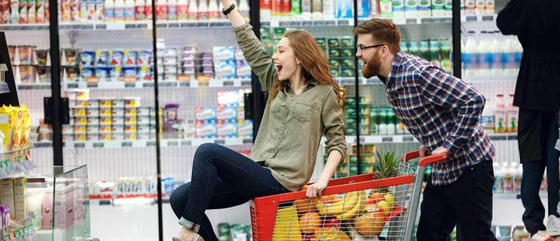 Man en vrouw rijden lachend met boodschappenkar door supermarkt